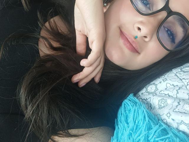 NataliaRivas with black hair, glasses, blue nose ring, and blue eyeshadow, lying on a blue fuzzy pillow, hand in hair, black shirt, smiling, fair skin.