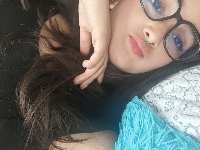 NataliaRivas with blue eyeshadow, black-framed glasses, and pouty pink lips, holding dark brown hair, next to a turquoise fuzzy pillow and white patterned pillow.