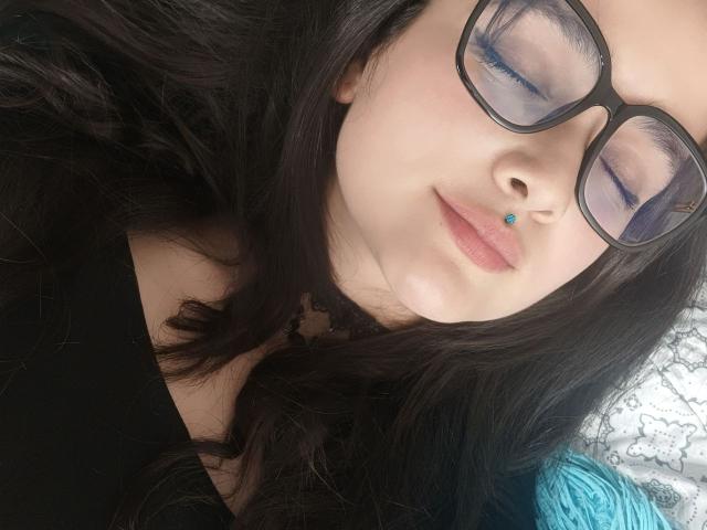 Close-up of NataliaRivas with dark brown wavy hair, wearing black-framed glasses, and a small blue nose piercing. Her eyes are closed with purple eyeshadow. She has fair skin and a slight smile. She's wearing a black top. The background includes a textured white and gray pattern, and a blue yarn is partially visible on the right. The image has a casual, intimate feel.