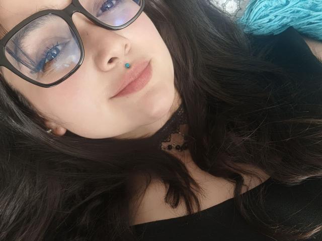 Close-up photo of NataliaRivas with long black hair, wearing black-framed glasses and a black choker. She has blue eyes, a blue nose piercing, and subtle makeup. She's wearing a black top with a low neckline. Her lips are slightly parted in a relaxed expression. The teal yarn in the background adds a subtle contrast. The composition focuses on her face, with the glasses and nose piercing drawing attention. Natural light highlights her skin's smooth texture.