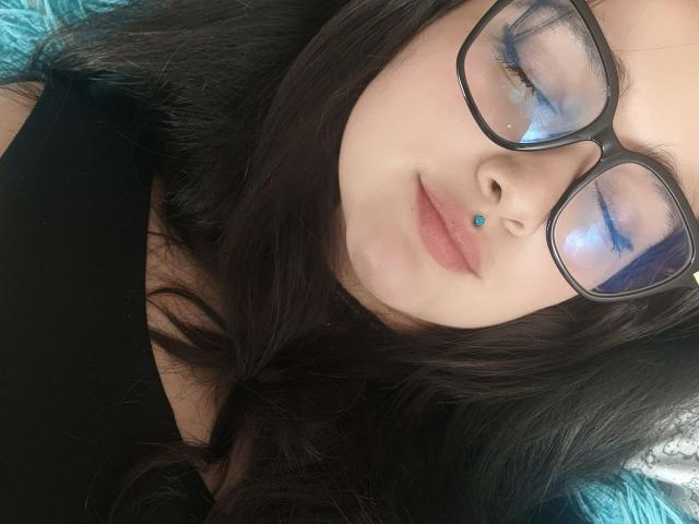 NataliaRivas with black glasses, blue eyeliner, nose piercing, and black hair, lying down, wearing a black top, eyes closed, slight smile, calm expression.