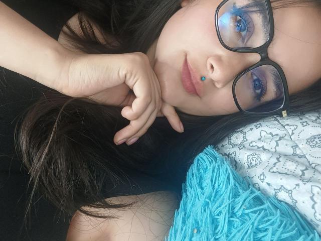 NataliaRivas lies on a bed, her face close to the camera. She has long dark hair, wears black glasses with a slight yellow accent, and has a blue lip piercing. Her right hand rests on her chin. She's wearing a black top and a blue, fuzzy pillow is to her right. Her makeup includes blue eyeshadow. The image is a close-up with soft, natural lighting.