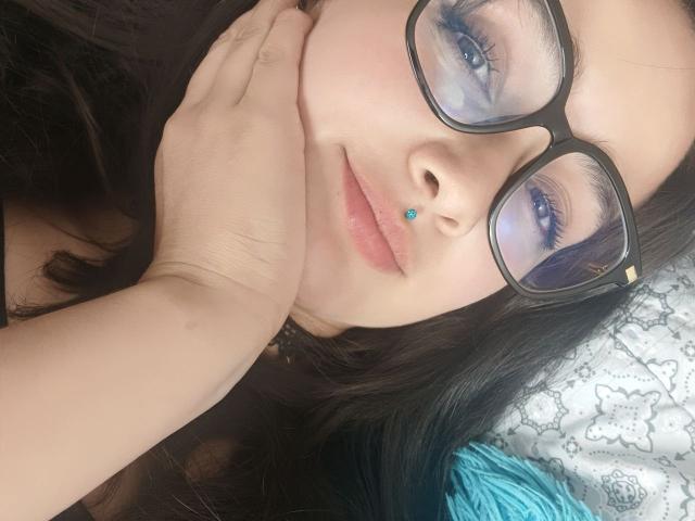 Close-up of NataliaRivas with dark hair, black glasses, and a blue nose stud. She rests her head on her hand, showing a slight smile. Her blue eyeliner and mascara contrast with her light skin. She has a blue ring on her finger. The texture of her hair is smooth, and the background includes a white, patterned pillow with teal accents. The image is focused on her face, with a soft, natural light.