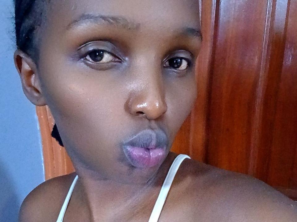 PrettyMercy with dark skin, pouty pink lips, and raised eyebrows, wearing a white strap, against a wooden door. Her eyes are slightly up, looking straight at the camera.