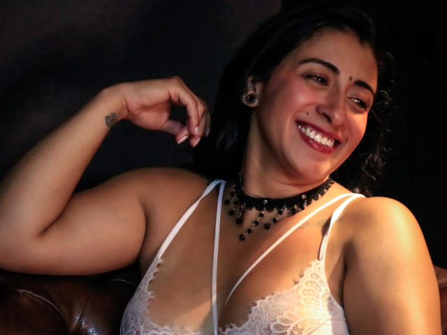 ZaraBeicker with dark hair, smiling, wearing white lace bra, black bead necklace, and black ear gauges, resting arm on brown leather couch. Her tits are small and cute.