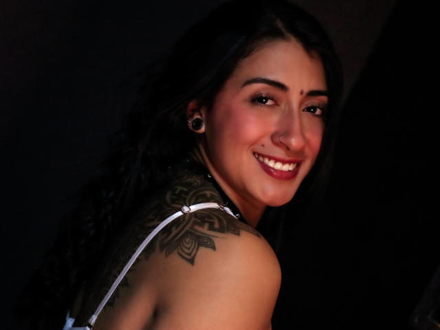 ZaraBeicker with dark wavy hair, smiling, showing off a tattoo on her right shoulder, wearing a white strap top, nose ring, and black ear gauges.