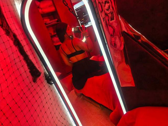 RouxiMori in a mirror, wearing a black bra and skirt, with a red neon light, sitting on a bed, her back to the camera, showing her ass.