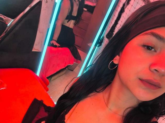 RouxiMori's face in the foreground with red lighting, showing her black hair, hoop earrings, and slight smile. Reflection in mirror shows her black dress and red bed.