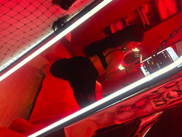 RouxiMori with long black hair, wearing a black bra and tight black shorts, kneeling on a red bed, taking a mirror selfie, showing her big ass and toned back.