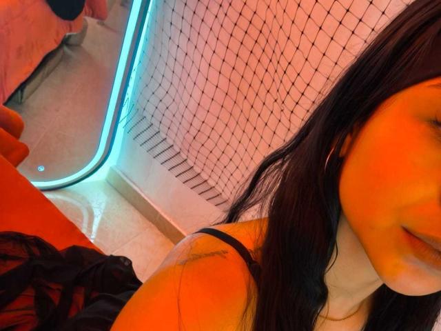 RouxiMori's face with orange lighting, black hair, and a subtle smile, wearing a black strap top, holding black clothes, and a blue-lit background.