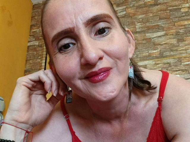 RubiAnderson has red lipstick, red bra, and stone wall background. She's smiling with pinkish skin, brown eyes, and wearing earrings with her hand near her mouth.