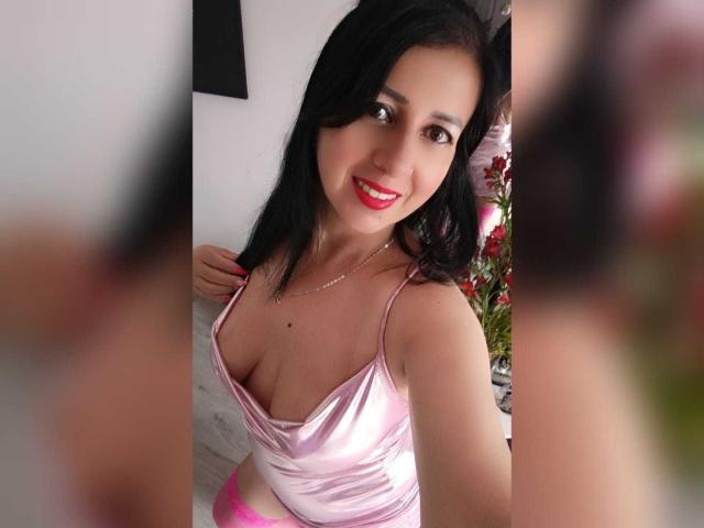 Briananice with dark hair, pink satin top, showing her small tits and pink underwear, smiling at the camera, red lipstick, silver necklace, mirror reflection, indoors.