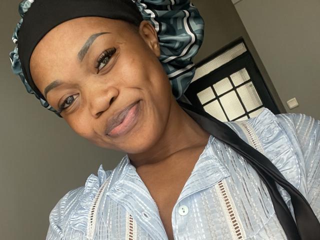 Sashie has dark brown skin, a black headwrap with green stripes, and a light blue patterned shirt. She's smiling with a slight pout, showing her pink lips.