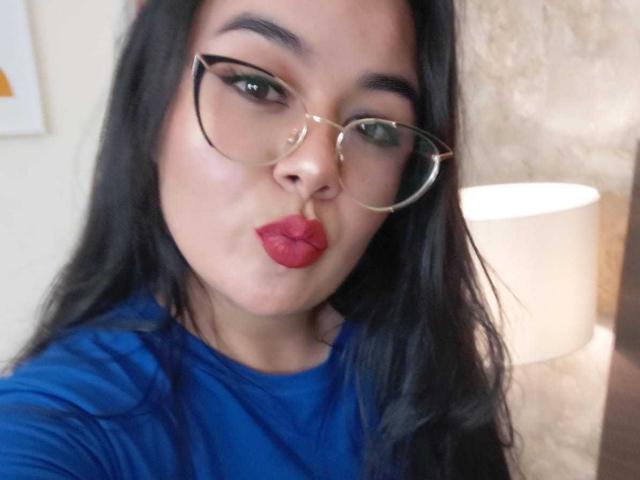 HannahMolly, com cabelos pretos longos, usando óculos e uma camisa azul, com lábios vermelhos curvados para baixo, olhos verdes e um piercing no nariz, em um selfie. Suas nádegas não estão visíveis..