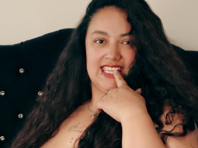AvaRyd with long black wavy hair, smiling, nose pierced, finger touching her lip, showing her teeth, tattoos on her chest, sitting on a black tufted couch.