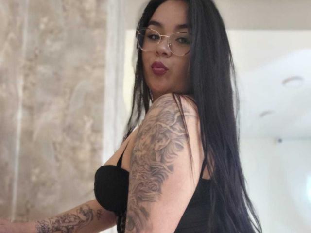 HannahMolly with long black hair, glasses, and red lipstick, wearing a black bra, showing off a detailed tattoo sleeve on her right arm. Boobs visible.