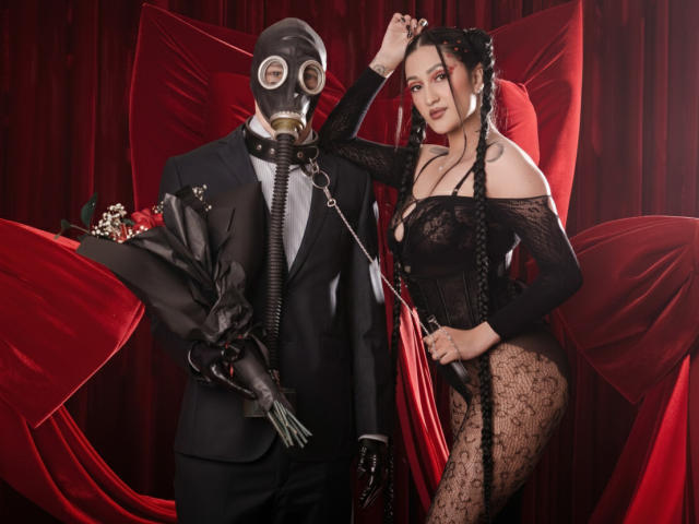 EmmaRosseti stands in front of red draped fabric, wearing a black lace bodysuit and black braids. She holds a black gas mask and leash, leading a man in a black suit with a gas mask covering his face. The man's suit has decorative flowers on the left lapel. EmmaRosseti's lace bodysuit is sheer, revealing her curvy figure and light skin. The man is also wearing black gloves. The composition is dark and dramatic, with red fabric creating a luxurious backdrop. The image is a photograph with a fetishistic theme.