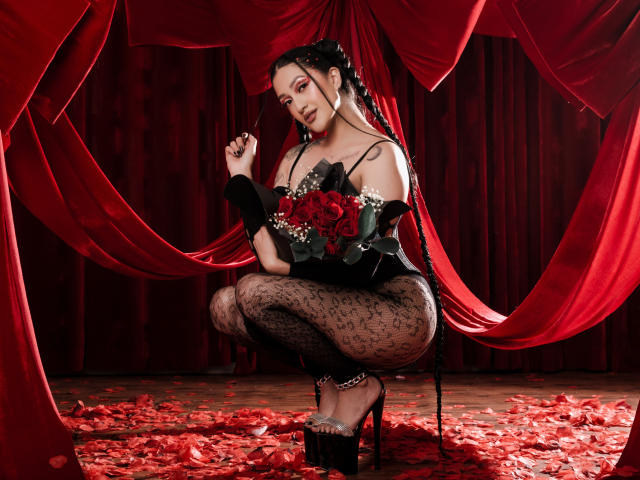 EmmaRosseti squats on a red-petal-covered stage, wearing a black lace off-shoulder top, sheer black lace pants, and silver high heels. She holds a bouquet of red roses with white petals. Red velvet curtains frame her, and her black hair is styled in braids with red accents. She wears hoop earrings and looks at the camera with a confident expression. The image has a dark, sultry vibe, with rich red and black colors.
