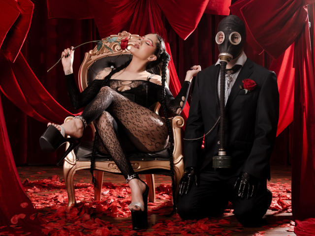 EmmaRosseti sits on an ornate, gold-trimmed chair, wearing a black lace bodysuit, high heels, and black stockings. She holds a cigarette, laughing with her head tilted back. Her dark hair is styled in a sleek updo. On the right, a man in a black suit, red rose in the lapel, and black gloves kneels with a gas mask covering his face, a cigarette holder in his hand. Red velvet drapes and scattered red rose petals fill the background. The composition is dark, dramatic, and sensual.