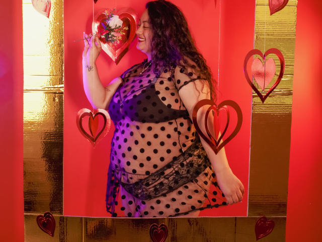 AvaRyd, a plus-sized woman with curly dark hair, stands in a vibrant red and gold setting. She's wearing a sheer black polka dot dress with lace detailing, exposing her large breasts and midsection. She holds a red heart-shaped decoration with flowers in her right hand, smiling. The background features gold vertical panels and red heart cutouts. The image uses bright, contrasting colors and highlights AvaRyd's confident, joyful presence.