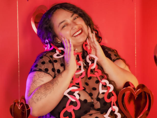 AvaRyd, a curvy woman with dark curly hair and a wide smile, sits against a bright red wall. She wears a sheer black polka dot top, revealing her large breasts, and a red and white twisted heart necklace drapes over her chest. Her hands are raised to her cheeks, showing a tattoo on her right arm. The image has a warm, red light cast on AvaRyd, with heart-shaped decorations on either side. Her expression is joyful and inviting.