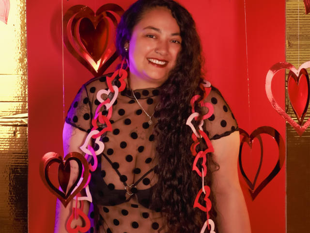 AvaRyd with long curly brown hair, smiling, wearing a sheer black polka dot top, red and white heart necklace, and a silver necklace, against a red background.