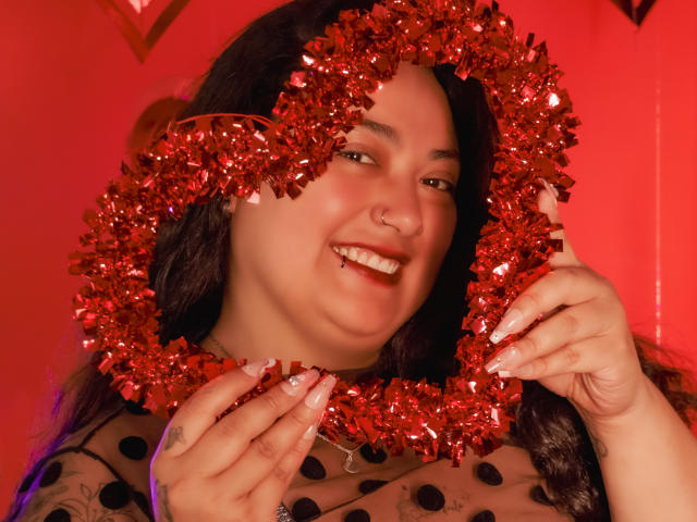 A portrait of AvaRyd with medium-length dark hair, wearing a red, shiny tinsel wreath around her head. She has a nose ring, is smiling with visible teeth, and holds the wreath with her fingers, showing long, manicured nails. She's wearing a sheer, polka-dotted top. The background is a solid red, and the lighting is warm, highlighting her skin and the tinsel's reflective quality. The image has a playful, festive vibe.
