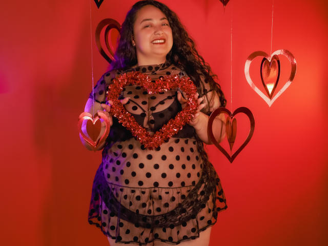 AvaRyd with curly dark hair, smiling, wearing a sheer black polka dot dress, holding a glittery red heart, and two wooden hearts against a red background.