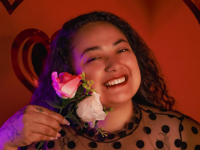 A close-up of AvaRyd with curly dark hair, holding a pink and white rose. She has a nose ring, a tooth gap, and a tattoo on her left hand. She wears a sheer black polka dot top. Her expression is happy with a wide smile. Warm red and purple lighting. The rose is in her right hand. The image has a heart-shaped element in the background.