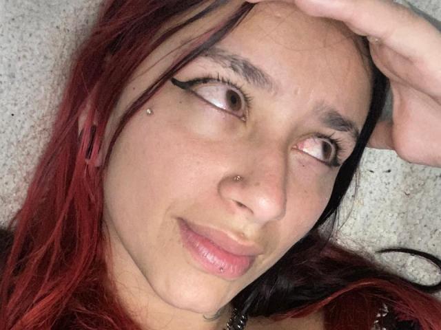 RoxanaSanchezz has red and black hair, brown eyes with black eyeliner, a nose piercing, a lip piercing, and a small facial stud, lying on a carpeted floor.