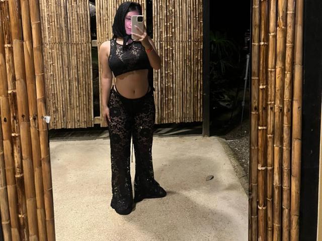 NicoleAnniston stands in front of a mirror, taking a selfie. She wears a black lace crop top and matching high-waisted, wide-legged pants. The lace has a floral pattern, revealing her midriff and parts of her skin. Her dark hair is wavy and loose. She holds a pink phone with a white case in her right hand. The bamboo wall and floor create a natural, rustic setting. The image is well-lit, highlighting the texture of the lace and her tan skin. The mirror's frame is bamboo, matching the wall.