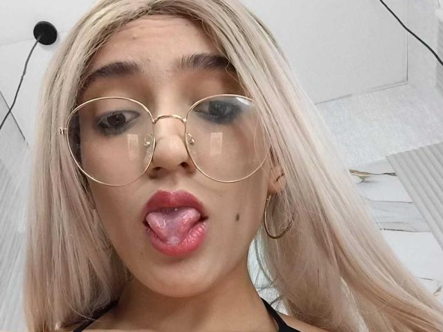 MichaelMistress with blonde hair, gold-rimmed glasses, and hoop earrings, sticking out her pink tongue, showing a small beauty mark on her left cheek.