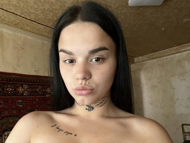Olivkaa has long black hair, a nose ring, and a tattoo under her chin with wings. She's topless, showing a tattoo on her right shoulder that says "a day in the sun".