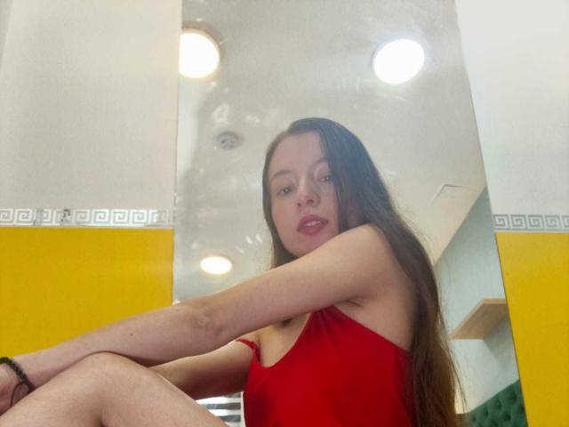 VioletaNova with long brown hair, red tank top, sitting, looking at camera, red lips, slight smile, yellow wall, green chair, arm bracelet, ceiling lights, mirror reflection.
