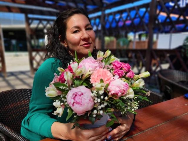 TinaMorel with curly black hair, green long-sleeve shirt, holding a pink and white flower bouquet, smiling, seated at a wooden table, wicker chair, outdoor setting.
