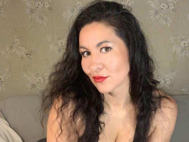 TinaMorel has wavy black hair, red lipstick, and brown eyes. She's looking at the camera with a slight smile, showing her tits in a low-cut top.