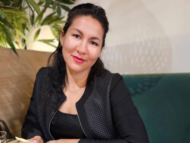 TinaMorel, a woman with medium skin tone and black hair, sits with a slight smile. She has bright red lipstick and wears a black, textured vest over a black top. Her arms are crossed. She has small gold earrings and a thin gold necklace. The background includes a green plant on the left and a dark green cushion on the right. The lighting is soft, creating subtle shadows. The image is a portrait photograph.
