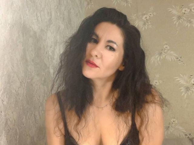 TinaMorel, a medium-skinned woman with wavy, dark brown hair, stands in the frame. She has red lipstick and subtle eye makeup. She wears a black spaghetti-strap top, showing cleavage. Her expression is slightly tilted, with a small, confident smile. A simple necklace rests on her chest. The image focuses on her upper body, with her hair partially covering her right eye. The background is a textured, beige wallpaper with floral patterns.