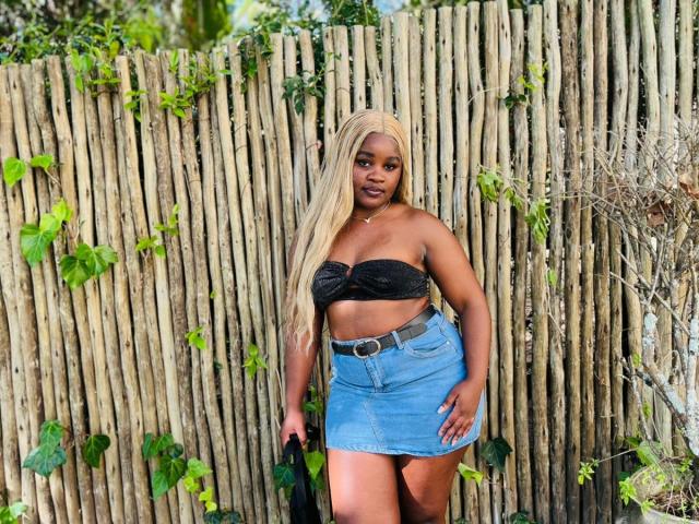 StellaJoanne stands in front of a wooden fence with green vines. She has long, straight blonde hair and a dark strapless top that ties in the center, revealing her midriff. She wears a blue denim skirt with a black belt and holds a black bag in her right hand. Her skin is dark, and she has a confident expression. The image has bright, natural lighting. Her body is curvy with an hourglass shape. The overall composition is casual and outdoor-focused.