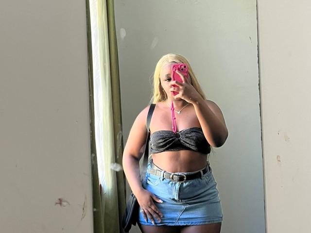 StellaJoanne with blonde hair, wearing a black strapless top, showing her tits, and a blue denim skirt, taking a mirror selfie with a pink phone, showing her ass.