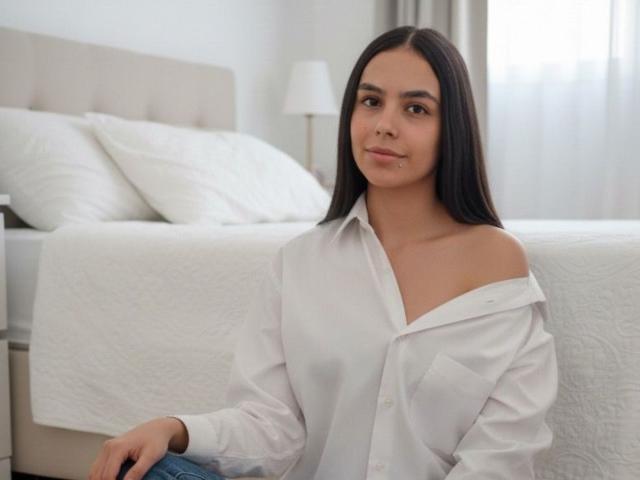 NaomiBlake with long black hair, wearing a white off-shoulder shirt, sitting in front of a bed with white pillows and a quilted blanket, looking at the camera.