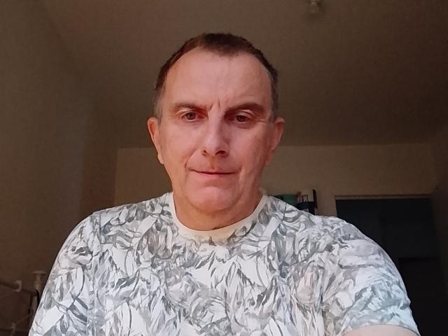 Belhomm6 is a middle-aged man with a receding hairline and short, dark brown hair. He has a slightly wrinkled forehead and a subtle, almost neutral smile. Belhomm6 is wearing a white and gray patterned shirt with a leafy design. The photograph is taken indoors with dim lighting. His skin is light and his eyes are slightly squinted. The background is blurred, focusing attention on Belhomm6. The image has a straightforward, realistic style.