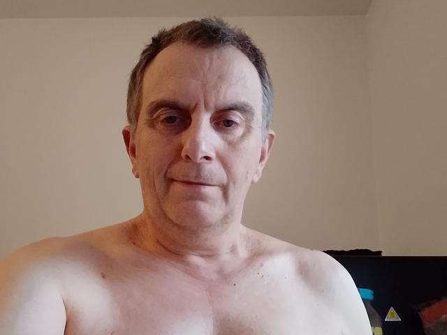 Belhomm69, shirtless, middle-aged man with short brown hair, slight wrinkles, pale skin, small ass, small tits, smiling, neutral expression, standing against plain background, visible TV on right.