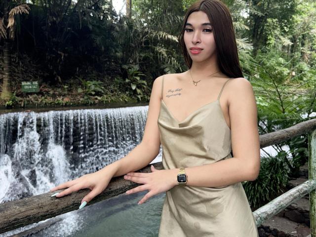Urhugecock-hot with long brown hair, tan dress, and gold watch, standing by a waterfall, hands on a wooden railing, tattoo on chest, smiling, green forest background.