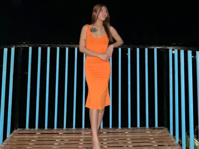 Urhugecock-hot stands on a wooden platform, wearing a bright orange, form-fitting dress that reaches mid-calf. She has long brown hair, a tattoo on her left shoulder, and is barefoot. Her right hand rests on her abdomen while her left hand is by her side. The background features a black fence with vertical blue stripes. The image is well-lit, highlighting her dress against the dark background.