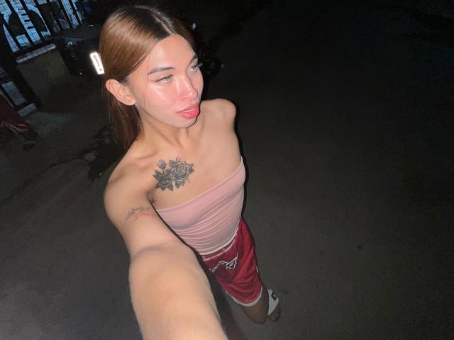 Urhugecock-hot with light brown hair, pink strapless top, rose tattoo on chest, red shorts, taking selfie, looking up, pink lips, dark background, visible arm tattoo.