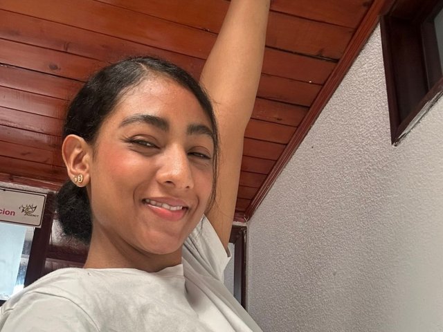 NatalyBlonder with dark hair, brown eyes, and a cute smile, raising her arm, wearing a white shirt, showing her small nose and tiny earrings, in a wooden ceiling room.