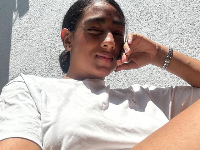 NatalyBlonder with dark skin, small nose, and earring, wearing a white tee, smiling slightly, hand near face, silver bracelet, sunlight, and shadow on textured wall.