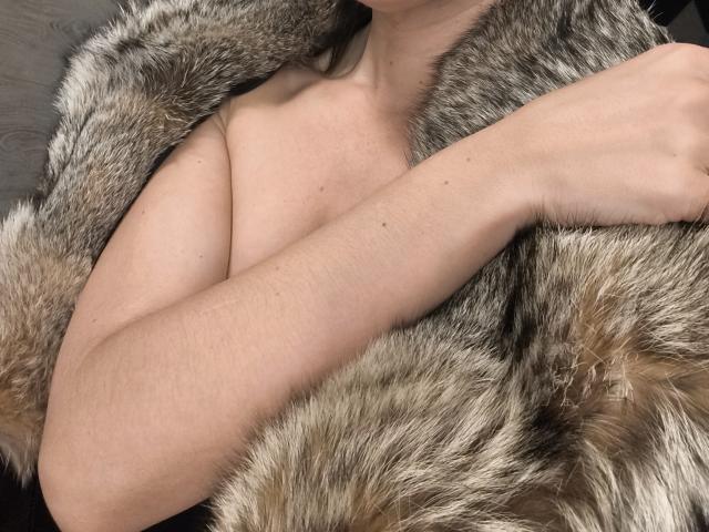 OrianaLaFrancaise with her arm crossed, wearing a fluffy grey and brown fur coat, exposing her bare, small to medium-sized tits. Her skin is light and smooth.