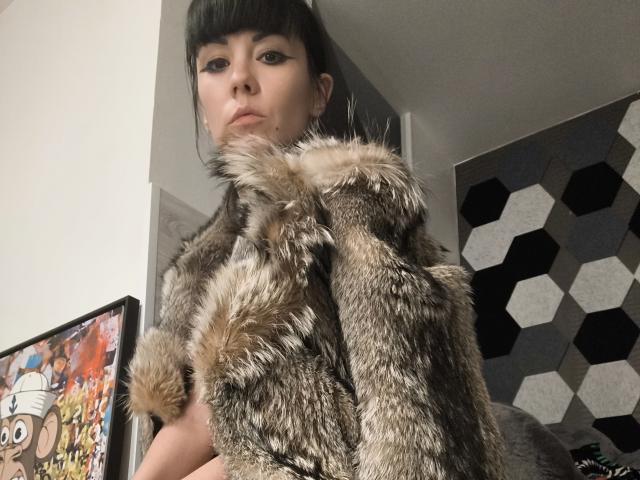 OrianaLaFrancaise stands in a room, wearing a brown, furry, fox-skin coat with prominent fur trim. She has dark, straight hair with bangs and heavy black eyeliner. Her expression is neutral, slightly pouty. The left side shows a colorful framed artwork with a cartoon character. The right background has hexagonal black and white patterns. The coat's texture is realistic, with detailed fur. OrianaLaFrancaise is centered in the frame.
